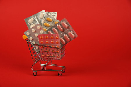 Close up several different blister packs of pills in small shopping cart over red background,...