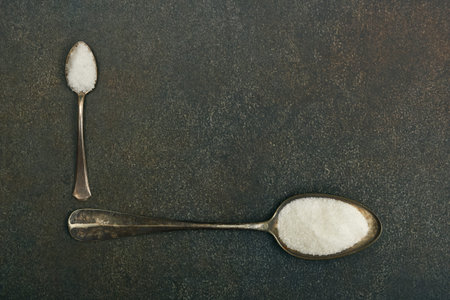 Close up flat lay two vintage metal spoons of white sugar and sea salt on dark grunge stone table...