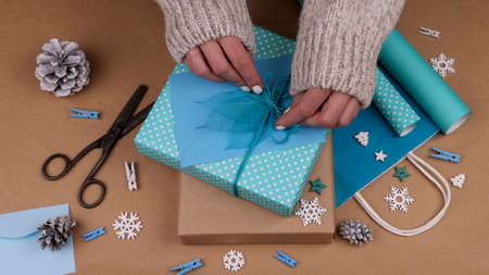 Close up woman hands packing and wrapping Christmas gift boxes with blue paper, adding envelope and skeleton leaves, high angle viewの写真素材