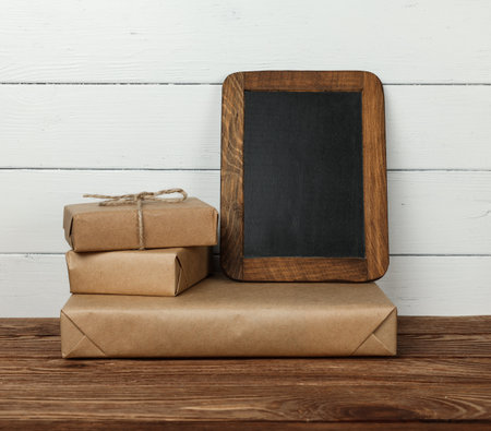 Close up stack of several brown paper wrapped gifts on wooden table and blank slate chalk board sign