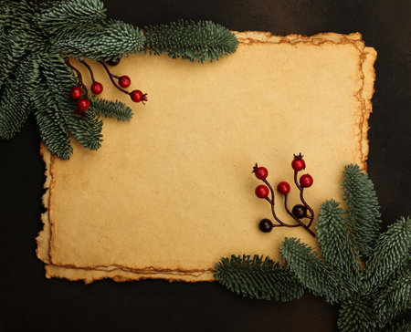 Close up several sheets of vintage paper parchment with Christmas decoration of fresh spruce branches, golden cones and red holly berries over dark brown background with copy spaceの写真素材