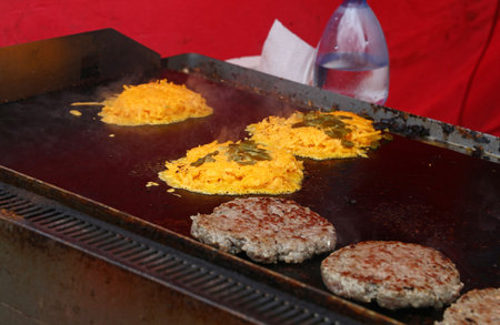 Ground beef patties and shredded cheese with jalapenos are sizzling on a large flat top grill, indicating the preparation of cheeseburgers.の写真素材