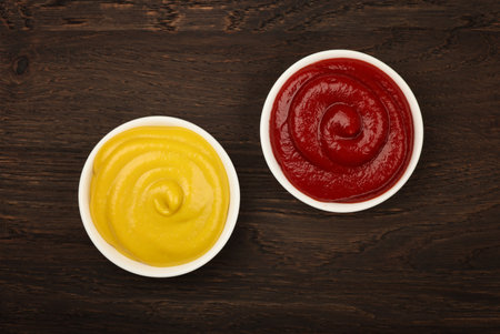Close up two white ceramic bowls of red ketchup tomato sauce and yellow mustard on brown table surface background with copy space, top view, directly aboveの写真素材