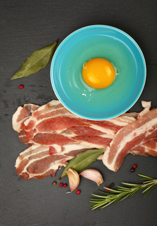 Fresh ingredients for a tasty breakfast, bacon, egg, garlic, herbs, and spices on a black slate,...