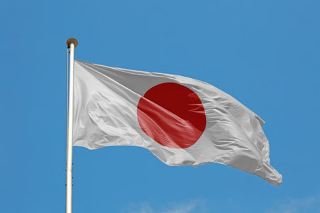 National flag of Japan flying and waving in the wind on flagstaff over clear blue sky, symbol of Japanese patriotism, low angle, side viewの写真素材