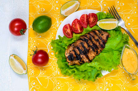 Fresh grilled chicken fillet with with lettuce, tomatoes and limes. Dish is in white wooden table on yellow cloth with fork. Ceasar sauce served together with dish.の写真素材