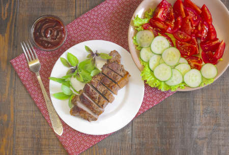 Grilled beef steak with salad and souce on wooden table at white plate. Salad with tomatoes and cucumbers and souce barbecue. Leafs of basil as decoration.の写真素材