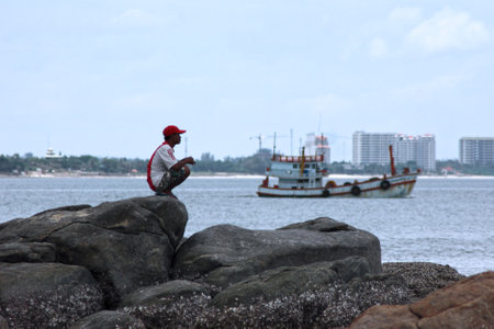 Hua Hin, Thailand - August 12 2007: Thai man squatting on a rock while looking at the horizon. Behind him, a fishing boat is going at sea.のeditorial素材