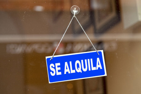 Blue sign hanging at the glass door of a shop saying in Spanish "Se alquila" meaning in English "To rent".の写真素材