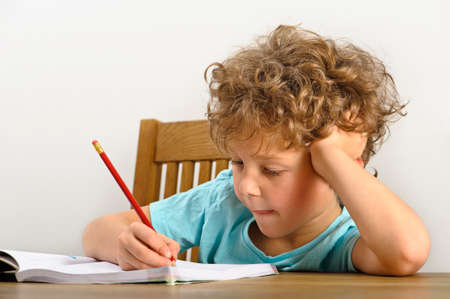 Curly haired young boy sits at a wooden table and does his homeworkの写真素材