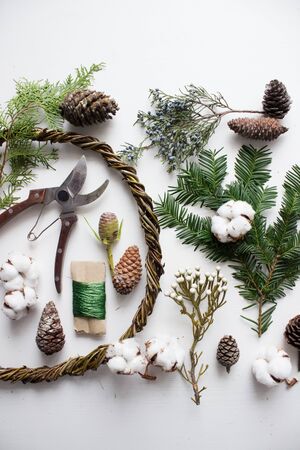 Top view on floristic selection for making rustic Christmas wreath - secateurs, basis of branches, wire, cones, cotton and pine branches on white tableの写真素材
