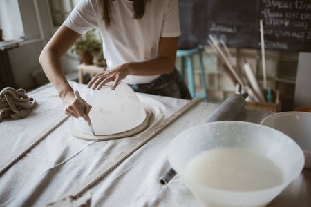 Sculptor cuts grey raw clay round plastic bowl for modeling ceramic plate on big table with industrial fabric in workshopの写真素材