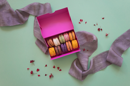 Colored fresh macaroons in pink box with silk lilac ribbon and little petals on light green backgroundの写真素材