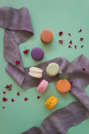 Colored fresh macaroons with silk lilac ribbon and little petals on light green background, top viewの写真素材
