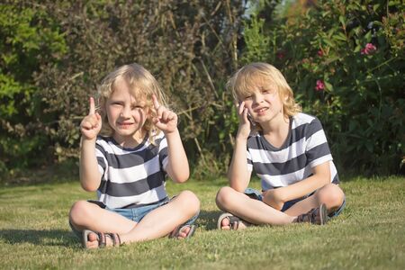 Two smiling blond boys are sitting on the lawn in the garden. One of them showed the fingers upward. The other is calling.の写真素材