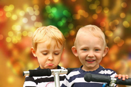 Pair of disappointed and happy blond boys in the striped shirts  on a scooters. Blurred Christmas background.のeditorial素材