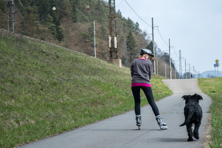 Middle aged woman is skating beside her dog outdoors. Rear view. の写真素材