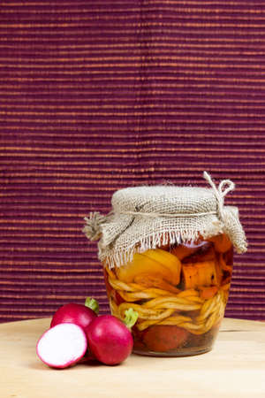 Jar of pickled variety of cheese in oil is standing on a wooden board. Reddish brown cloth background. Two whole and one halved radishes are lying next to the jar.の写真素材