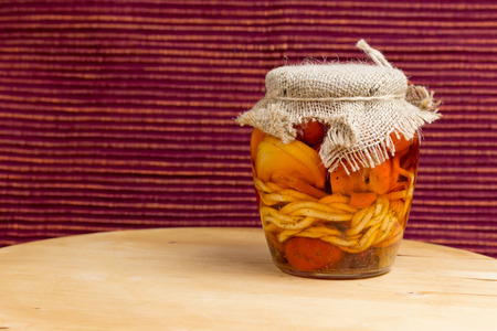 Jar of pickled variety of cheese in oil is standing on a wooden board. Reddish brown cloth background.の写真素材