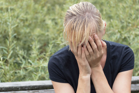 Desperate blonde middle aged woman in black shirt is sitting on the bench outside. Hands are covering her face.の写真素材