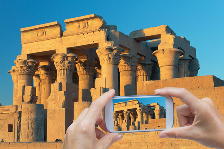 In the right bottom of the photo are hands holding smart phone and taking picture of Kom Ombo Temple dedicated to Sobek the crocodile god, and Horus the falcon-headed god (Egypt)の写真素材
