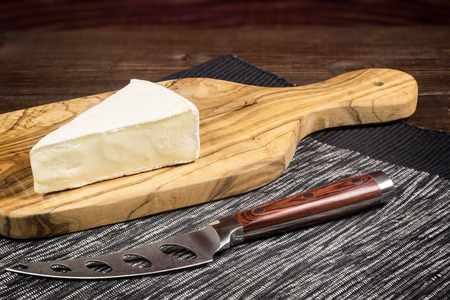 Cheese with white mold it is lying on the board of olive wood. In a front of the cheese  on a tablecloth is lying stainless steel cheese knife with wood handle. Photo is edited as an vintage with dark edges.の写真素材