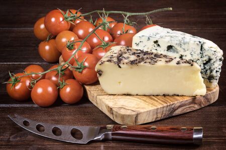 Two kinds of cheese and cherry tomatoes are lying on the board of olive wood. Stainless steel cheese knife with wood handle is lying on a wooden board in foreground. Photo is edited as an vintage with dark edges.の写真素材