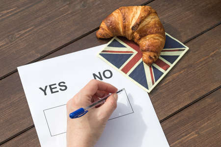 Aerial view of a female hand is ready to fill NO in the printed form. On the wooden table is lying a napkin English flag design, on which is symbolically lying a French croissant.の写真素材