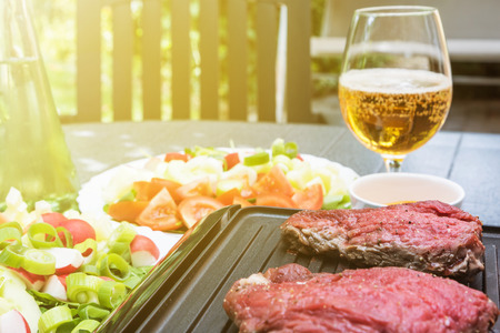 Grill steaks are on an electric stove. Bottle with oil, various kinds of vegetables and glass of beer are standing on the table in the sunshine.の写真素材