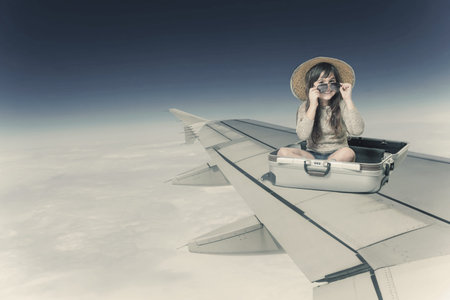 Smiling little girl in glasses and hat is sitting in the open suitcase on the wing of the plane in flight. Edited as a vintage photo.の写真素材