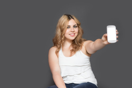 Studio shot  of attractive long haired smiling teenage girl showing smart phone with  blank touchscreen. All is on the gray background.の写真素材