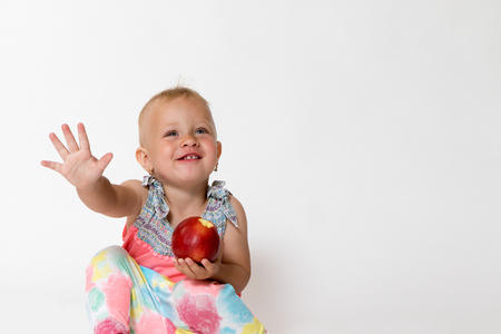 Studio shot of smiling toddler girl is sitting, gesticulating and holding red appleの写真素材