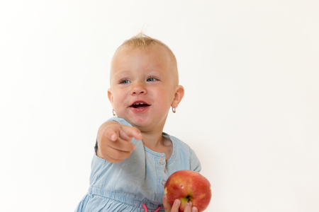 Studio shot of sitting cool toddler girl holding red apple and pointing her finger at the cameraの写真素材