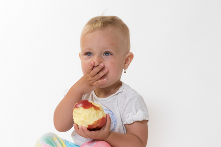 Studio shot of sitting cool toddler girl holding red apple and covering her mouth with handの写真素材