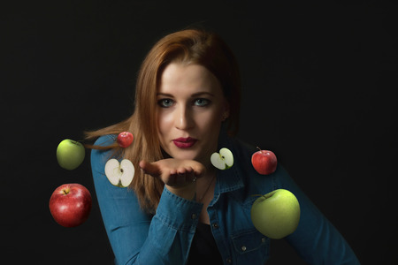 Low key portrait of sensual young woman sending a kiss with a open palm. Red and green apples are flying around her.の写真素材