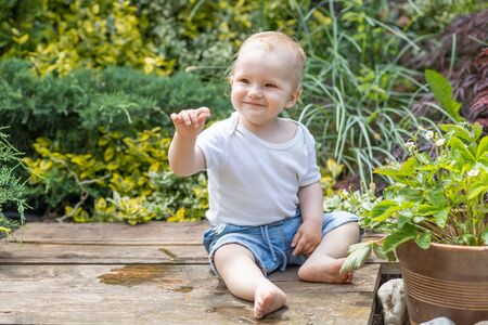 Smiling little baby boy is sitting next to a flowerpot with plants in the gardenの写真素材