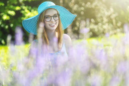 Portrait of smiling young woman with glasses wearing turquoise summer hat is posing behind lavender in the gardenの写真素材