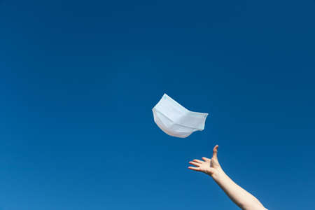 Symbol of the abolition of the obligation to wear a protective face mask. The female hand is throwing a protective face mask against blue sky. There is enough free space for your text in the photo.の写真素材
