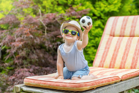 Little baby boy is sitting on the deck chair in the garden showing little soccer balloon.
