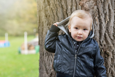 Adorable little boy dressed  in a leather jacket is taking off his hood outdoors.
