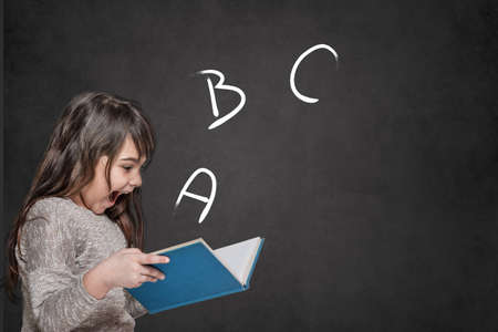 Cute tanned long haired  little girl is surprised at the letters ABC coming out of an open book she is holding in her hands.の写真素材