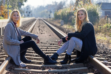 Two attractive young women  are sitting on rail in autumn backlight. Horizontally.