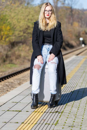 Young woman is sitting on suitcase in the railway station looking at the camera in the sunny autumn dayの写真素材