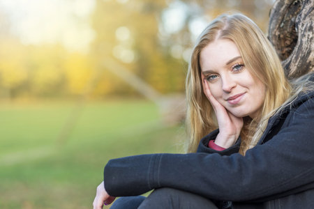 Smiling young woman is posing in the autumn park supporting her head with  hand looking at the camera. Horizontally.の写真素材