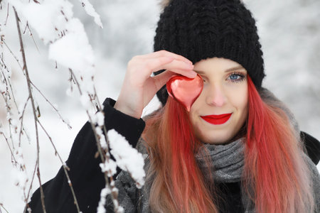 Front view portrait of a attractive red hair girl covering one eye with a red heart in snowy winter day.の写真素材