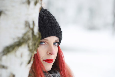Portrait of red hair girl ibehind a birch tree. The girl is looking at the camera.