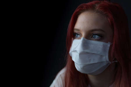 Closeup low key portrait of young redhead woman at home on quarantine with medical mask. Woman is looking at the side.の写真素材