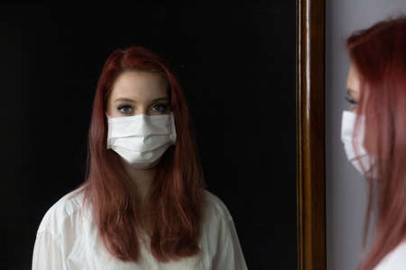 Young woman with medical mask is looking at herself in the old mirror. Horizontally.の写真素材