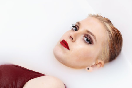 Attractive young woman with blue eyes and red lips lying in spa bath with milk and looking at...