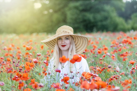 Portrait of blonde young woman in straw hat posing in red poppy field smiling at the camera. Horizontally.の写真素材